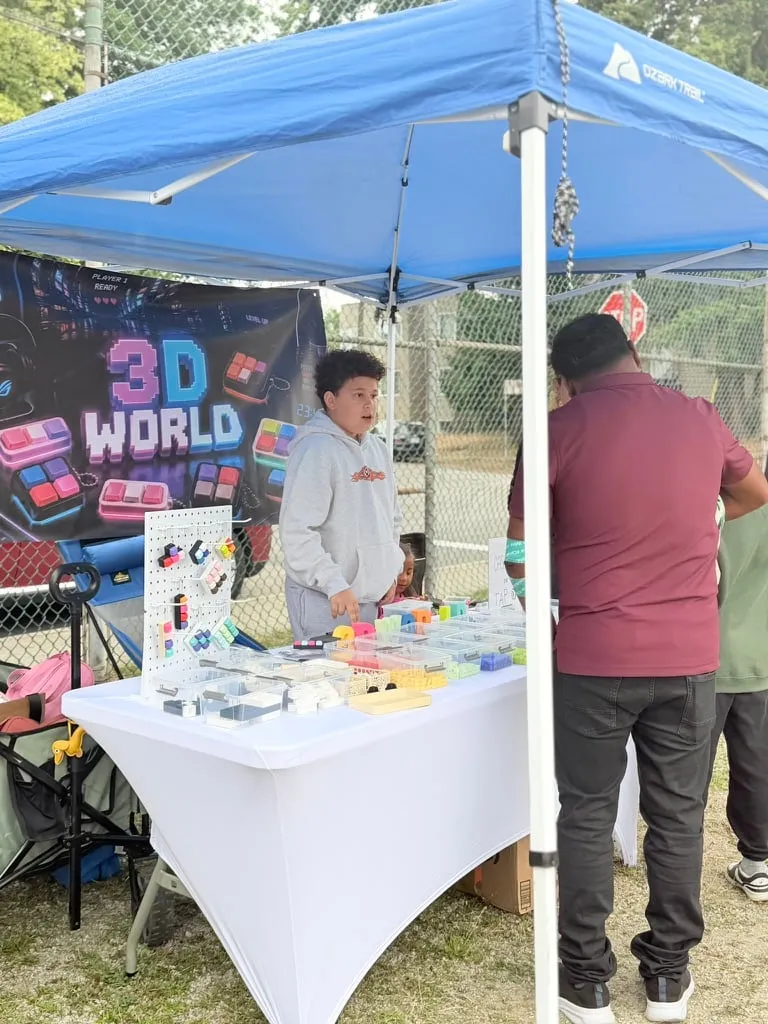 A young entrepreneur talks with customers at a booth displaying colourful 3D-printed objects.