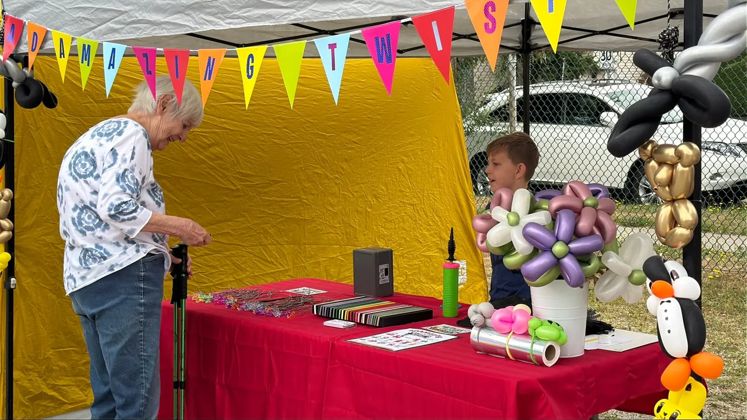 A young entrepreneur at a balloon-art booth talking with a customer across a table of balloon flowers and animals.
