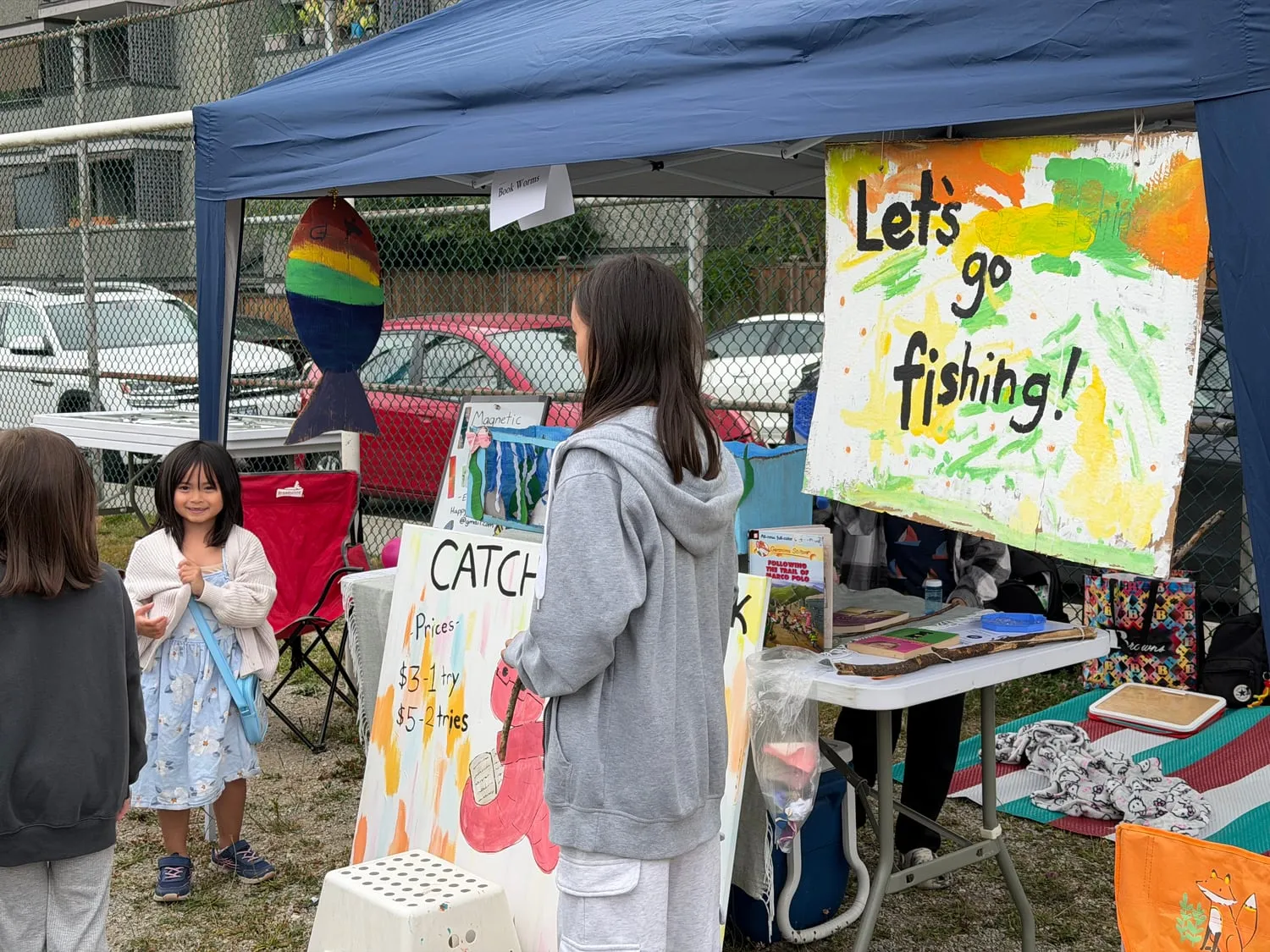 The Book Worms booth under a blue canopy, with a hand-painted "Let's go fishing!" sign and a price list for its magnetic-bookmark game.
