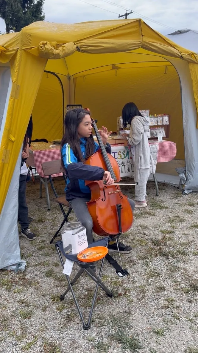 A young entrepreneur plays a cello in front of a yellow market tent while another child stands at a booth behind her.