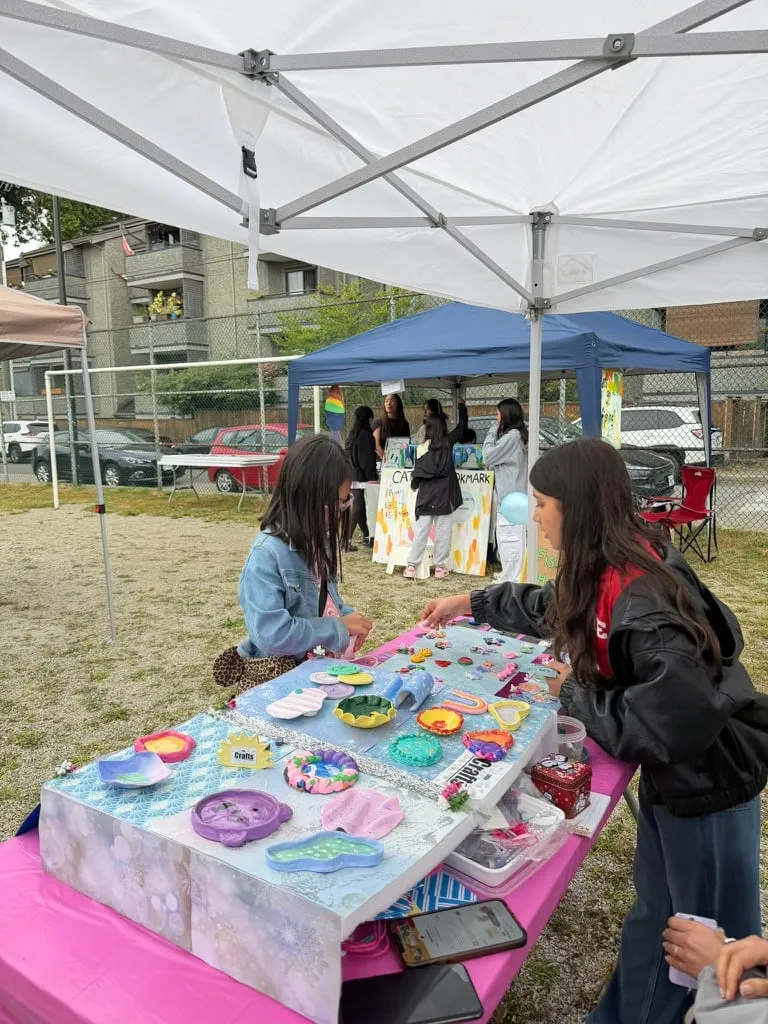 Two children talk across a table covered with colourful handmade crafts beneath outdoor market tents.